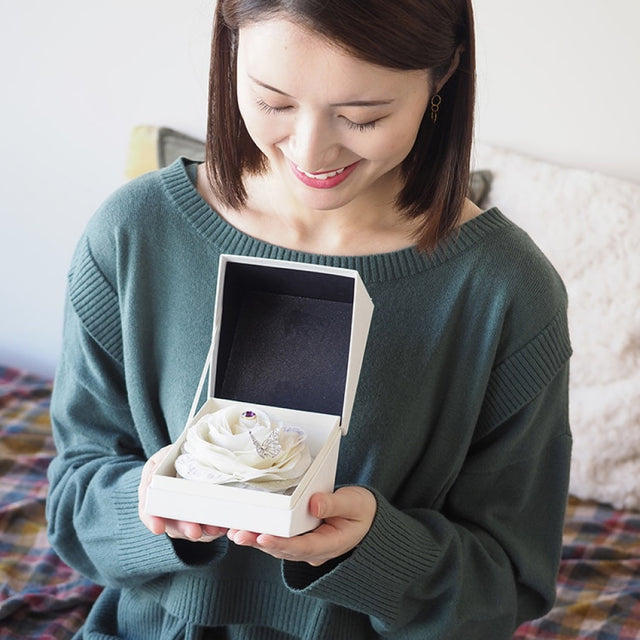 誕生石と刺繍が入った白バラのフラワーギフトを受け取り、嬉しそうに微笑む女性。部屋でプレゼントを開ける特別な瞬間を演出。