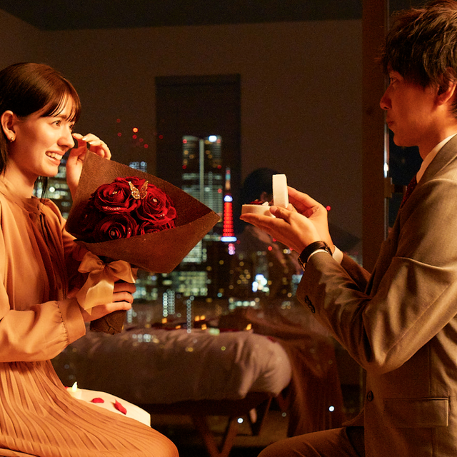 夜景の見えるホテルで、彼女にプロポーズをする男性。彼女は赤いバラの花束を抱え、男性は指輪の代わりになる特別なフラワーギフトを手渡している。