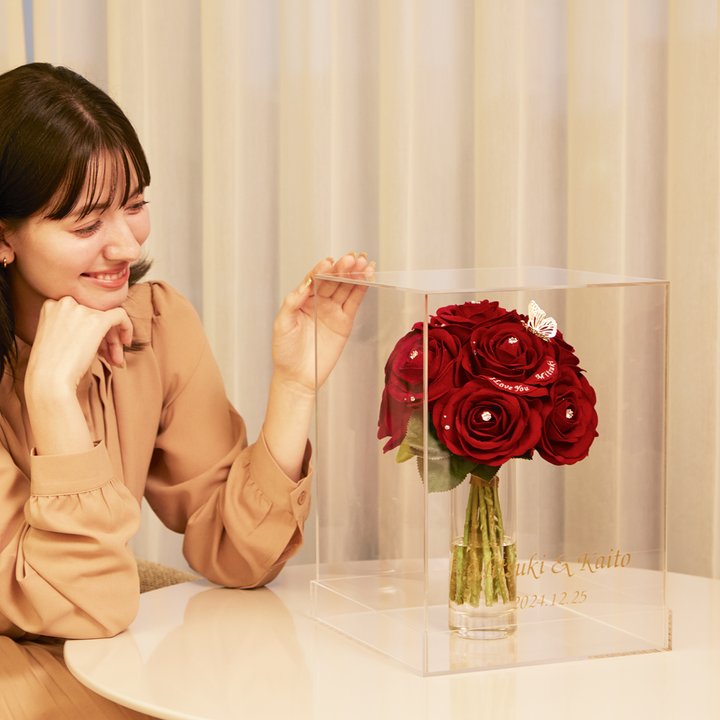 花瓶に入った12本の赤いバラが、透明な四角いケースに収納されている。ケースに入れると花が汚れず、美しいまま保管できる。ケースには二人の名前や記念日を入れられるので、特別感がある。