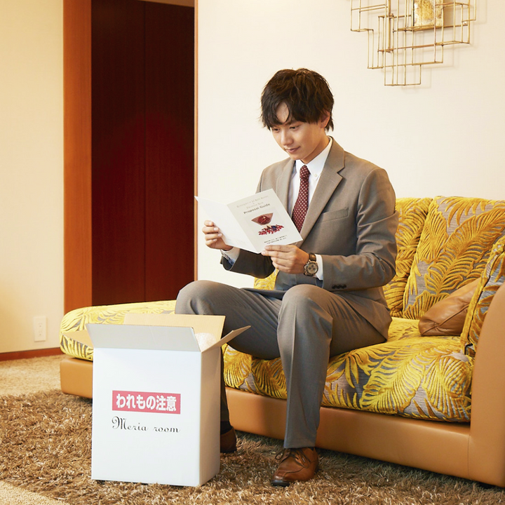 ホテルで届いた商品を開封し、手順書を確認している男性。プロポーズを成功に導く手順書が、商品を一緒に同封されている。