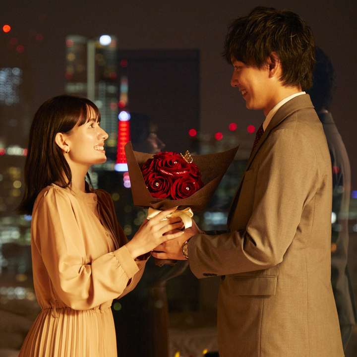 夜景の見える特別な場所で、赤いバラの花束を手渡しながらプロポーズをしている男性。プロポーズ場所として、夜景やホテルはロマンチックで人気。