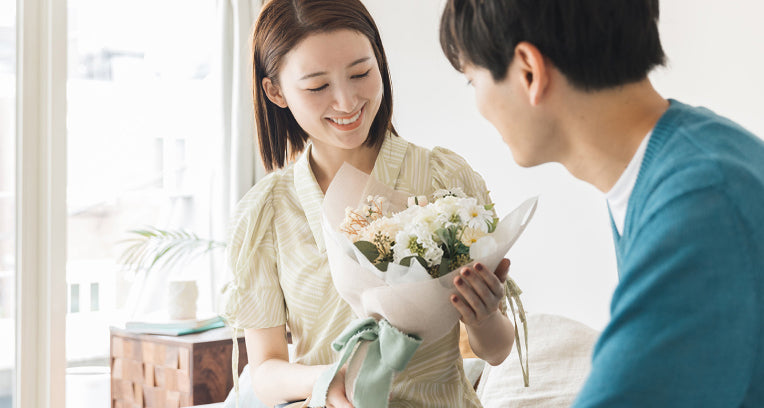 ホワイトダズンフラワーを見つめて微笑む女性と、それを見守る男性の姿。白を基調にしたナチュラルな12本の花束は、プロポーズや誕生日、記念日の贈り物にぴったり。部屋で贈るサプライズシーンにもおすすめの、メッセージ入り刺繍や花言葉カード付きギフト。