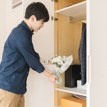 サプライズの準備中。男性が笑顔で花束をクローゼットに隠す、ワクワク感あふれるプロポーズ前のひととき。ナチュラルなホワイトダズンフラワーで、感動の瞬間を演出。