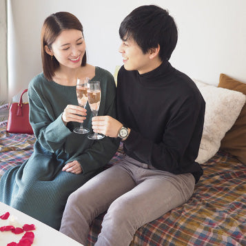 家でプロポーズを終えたカップルが笑顔で乾杯している様子。温かく幸せな空気に包まれた記念日のワンシーン。テーブルには赤い花びらとキャンドルの演出セットが置いてあり、特別な空間を演出している。