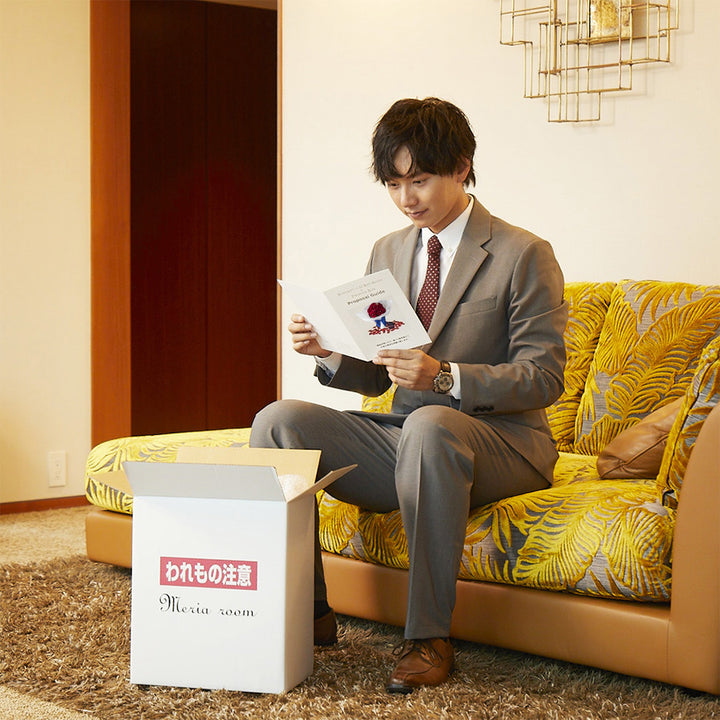 ホテルで届いた商品を開封し、手順書を確認している男性。プロポーズを成功に導く手順書が、商品を一緒に同封されている。