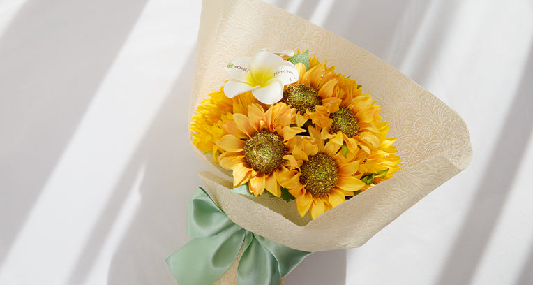 夏の代表花・ひまわりを贅沢に束ねた『ひまわりの花束とガラス花瓶セット』。白いプルメリアに名入れもできる特別仕様で、食卓やリビングに映える爽やかなフラワーギフト。誕生日やプロポーズなど大切な贈り物におすすめ。