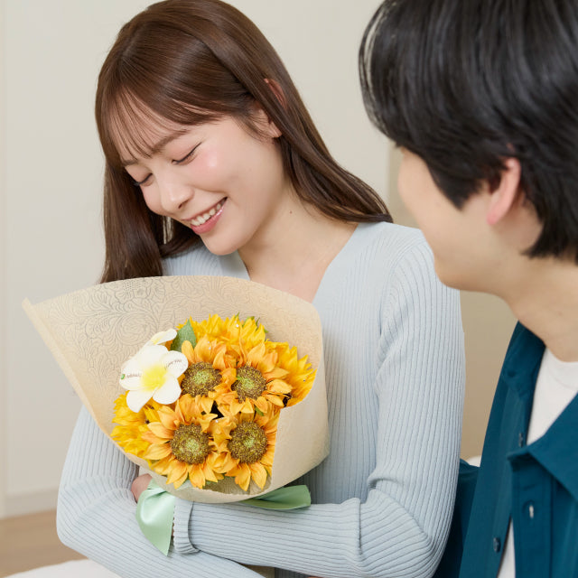 ひまわりの花束とガラス花瓶セットを手に、笑顔を見せる女性。夏らしいビタミンカラーのひまわりと白いプルメリアが映える、誕生日やプロポーズにぴったりなフラワーギフト。