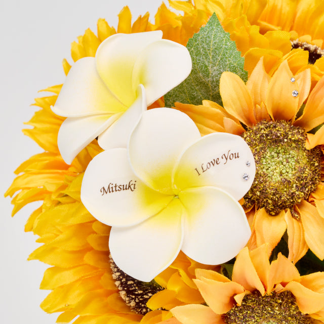 ひまわりの花束に添えられた白いプルメリアの花びらに、名前とメッセージを入れられる特別仕様。夏らしいビタミンカラーと爽やかさを兼ね備えたプロポーズや記念日のフラワーギフト。