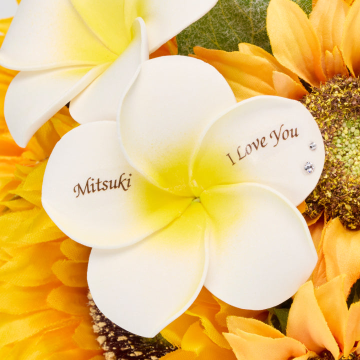 白いプルメリアに名前と愛のメッセージが刻まれた特別なひまわりの花束。名入れできるフラワーギフトは、プロポーズや誕生日など特別な日の贈り物にぴったり。